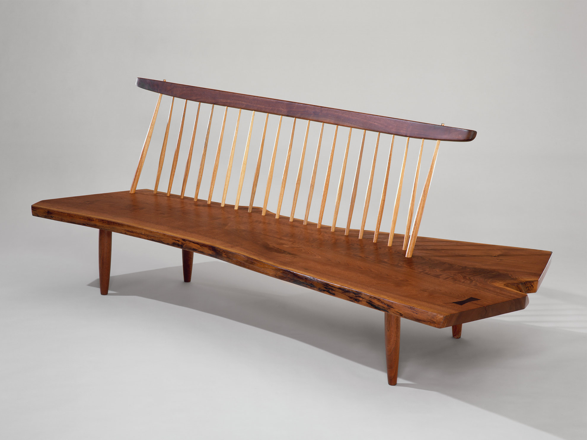 A slim walnut plank sits atop 21 bowed hickory dowels, which are affixed to a cross section of a dark walnut tree to form a bench. Four tapered walnut cylinders descend from the plank’s underside.