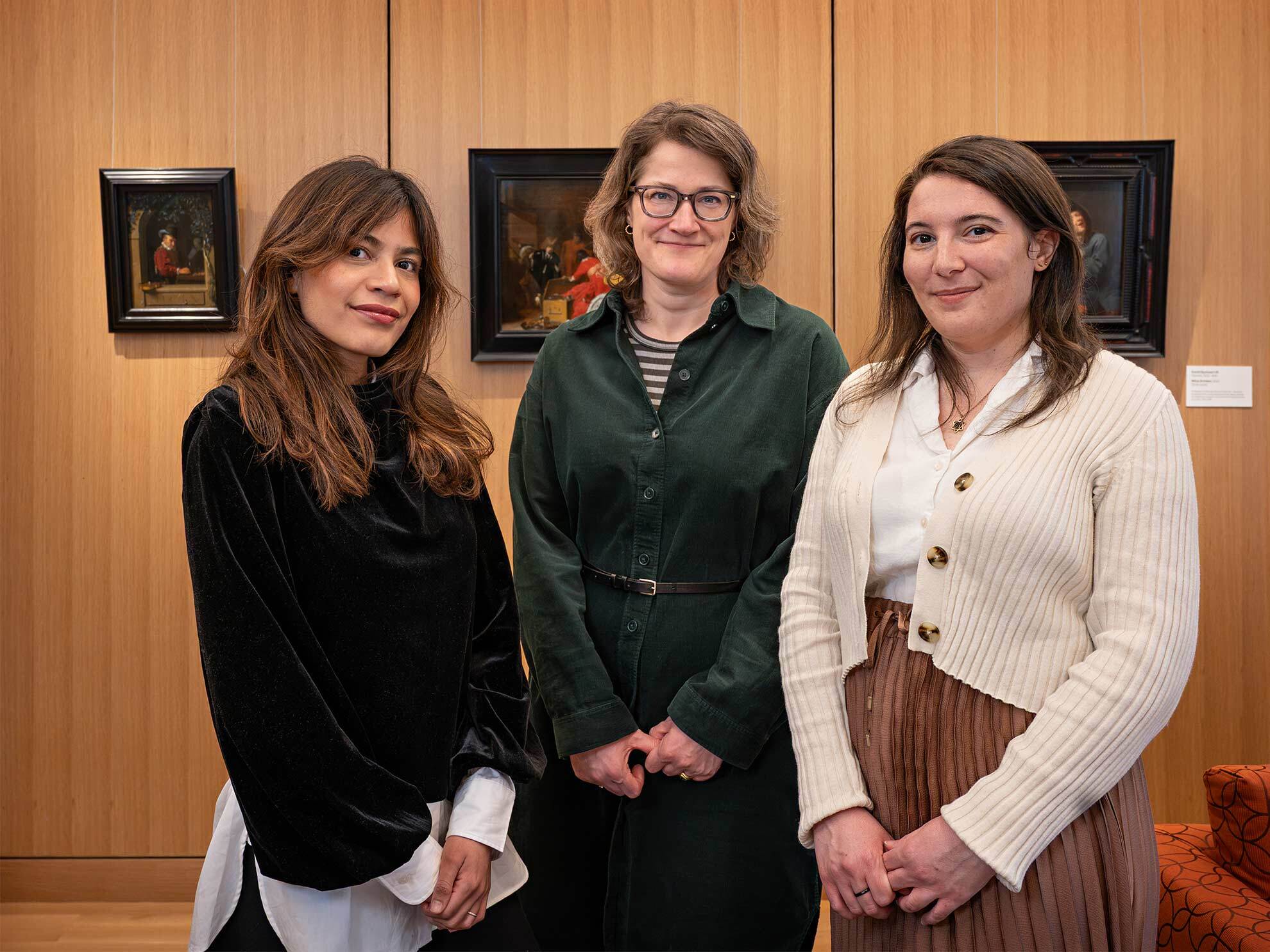 Three people stand in front of a wall with three framed paintings hanging.