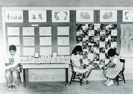 Children in a classroom sew names onto a quilt.