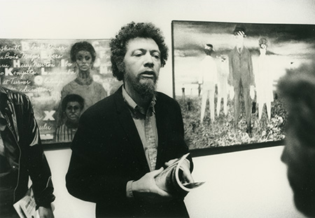 A bearded man stands in front of artworks on a wall.