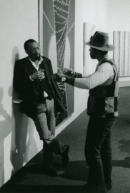 Two men stand and have a conversation in front of artwork in a gallery.