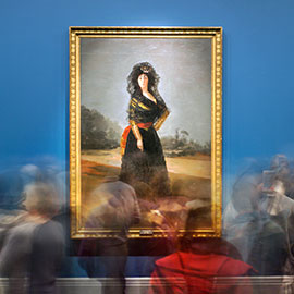 Motion-blurred visitors standing in front of Goya portrait of woman in black veil and dress with red sash around waist