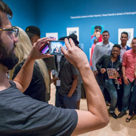 Visitor taking photo of group posing in gallery with prints hanging on blue wall