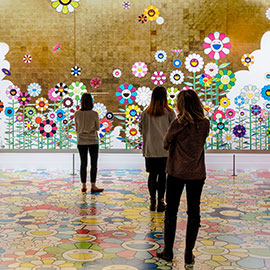 Three visitors standing in front of large Murakami mural with brightly colored flowers against tiled gold background