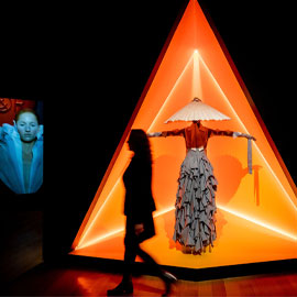 Visitor walking past multi-layered dress with conical hat displayed in bright orange platform