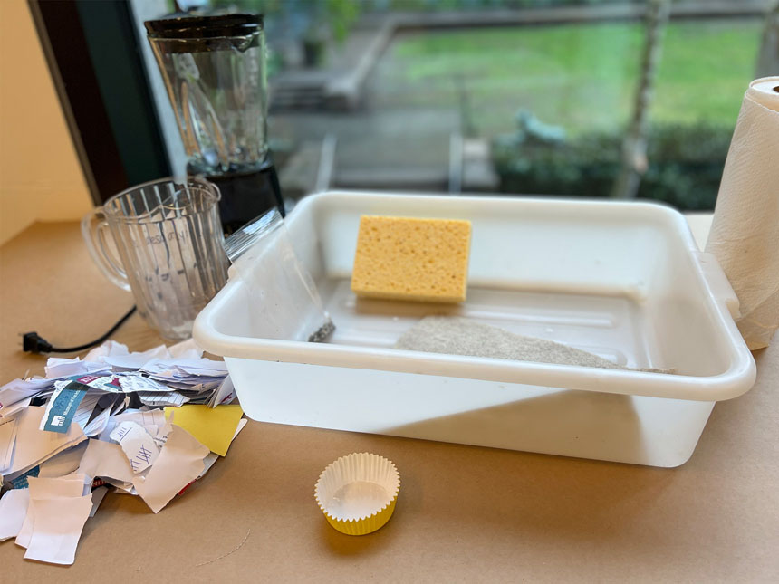 arrangement of materials used for the project laid out on a table: pitcher, blender, plastic bin, shredded paper, sponge