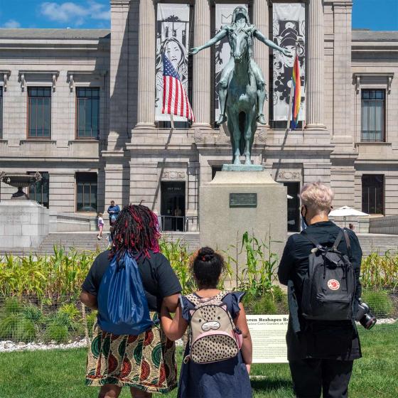 Three people stand with their backs to the camera looking at a sculpture of a Native American man on horseback.