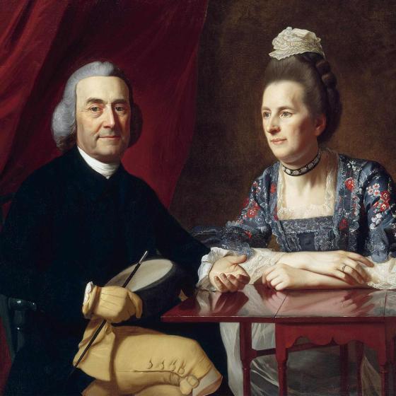 Before a lush red fabric curtain, a man in a powdered wig sits at a mahogany table, next to a woman in a dress with an elaborate floral pattern and a small cap.