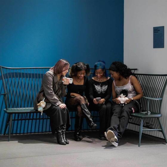 Four people sit on a dark green curved wooden bench in a corner of a room with blue and white walls.