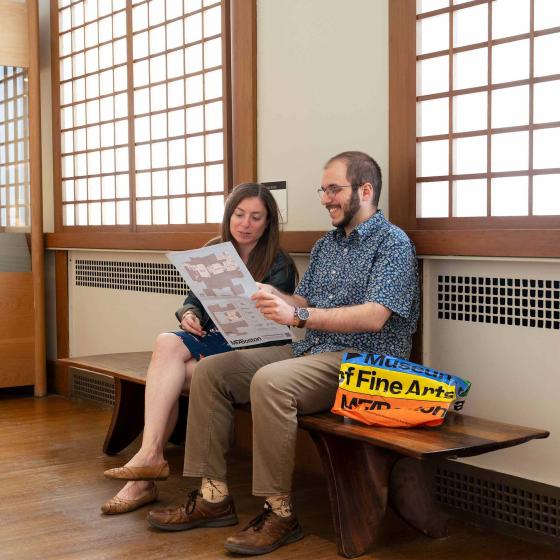 Two people sit on an orange and deep brown wooden bench in a brightly lit hallway.