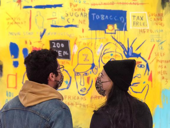 Two visitors wearing masks standing side-by-side and looking at the brightly colored Basquiat mural