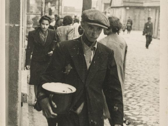 historical black-and-white photograph depicting young Jewish man in coat and jacket walking down sidewalk carrying a large bowl