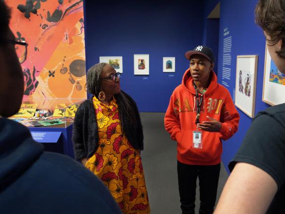 Teen speaking in front of small group, standing next to artist Ekua Holmes, in Paper Stories exhibition