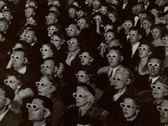 A photograph of a large audience transfixed by a movie; each audience member wears a pair of 3-D glasses