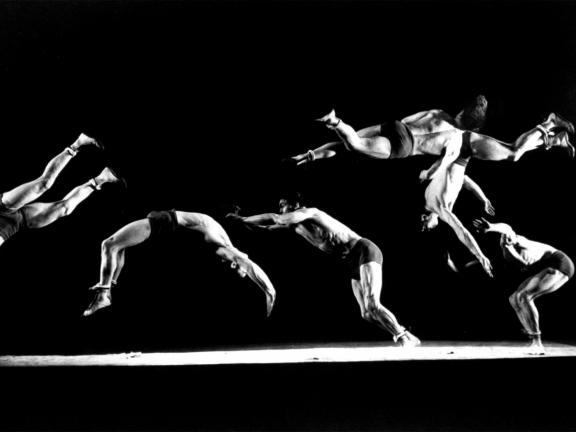 A black and white, stroboscopic photograph of gymnast New Loken tumbling and flipping across a gymnastics floor