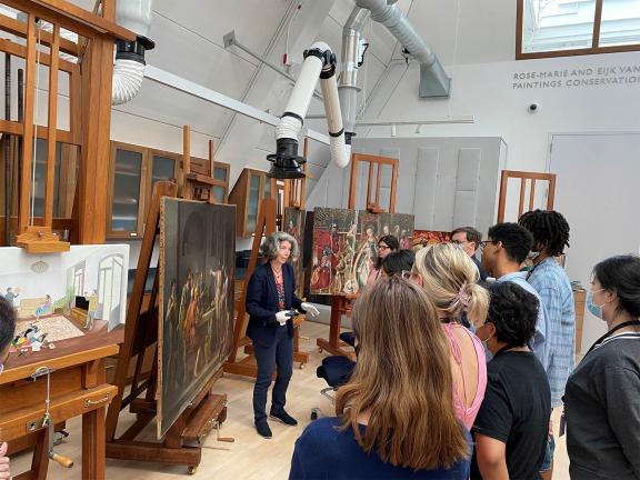 An MFA Conservator stands amongst multiple paintings on easels and addresses a small crowd.