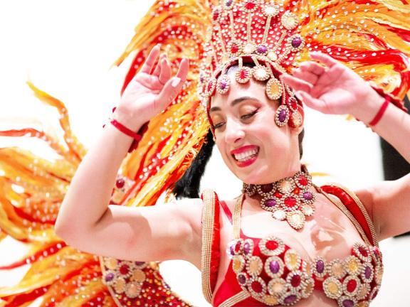 A woman smiling and dancing in a colorful, embellished outfit and feathered headpiece.