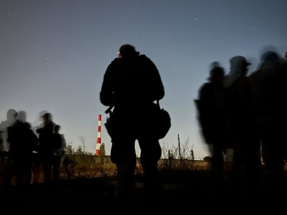 The silhouettes of Ukrainian soldiers stand out against the evening sky; a fluorescent orange and white tower sits in the distance.