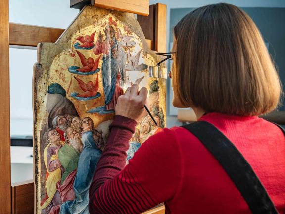 Conservator gently using thin brush on painting on wood panel depicting apostles looking up at Christ surrounded by angels