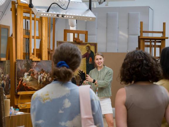 A speaker in a conservation lab talks with a small group of people in front of European artworks