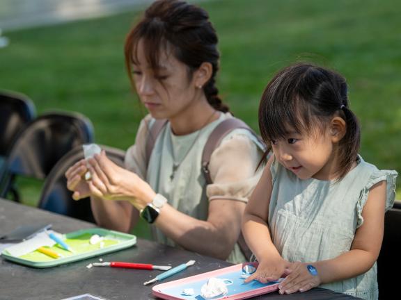 An adult and a child sit at a table outdoors on the Huntington Avenue Entrance lawn and work on an art activity