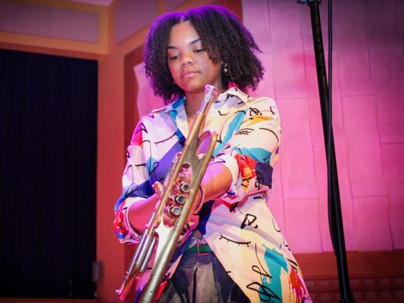 Colorful photo of a young woman in a graphic top with a trumpet in her hands and looks to be getting ready to play