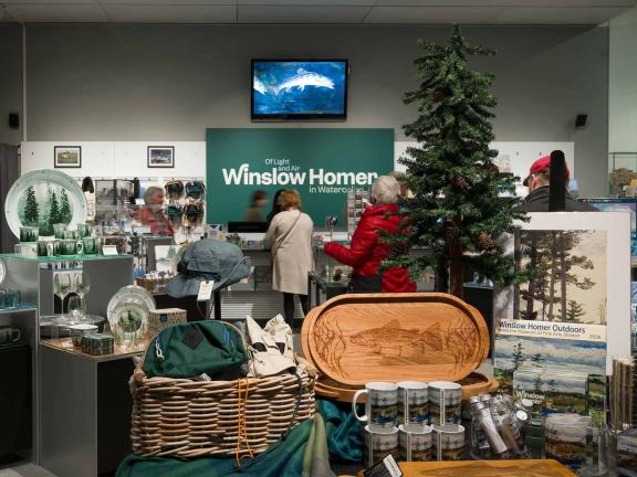 View of Winslow Homer gift shop with various nature-themed mugs, plates, bags, and calendars in foreground, in front of small evergreen tree; visitors shopping in background