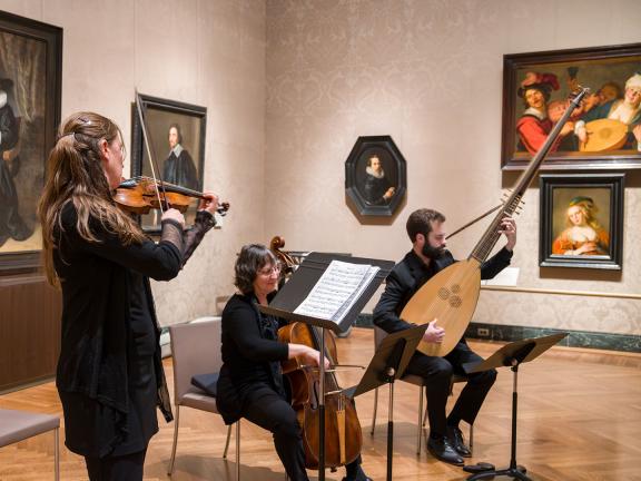 Three musicians playing various stringed instruments in gallery with Dutch paintings in background