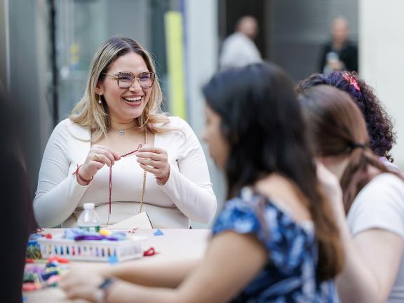 A woman smiles as she breads two threads—one red and one black—together.