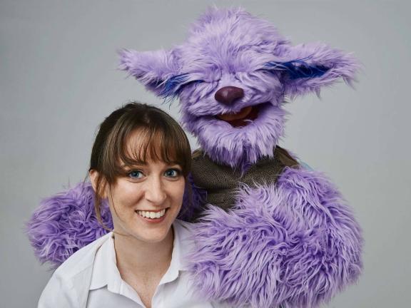 A light purple, fuzzy puppet with large pointy ears places its hand on a smiling woman's shoulders