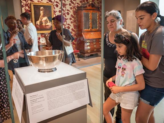A woman and two children stand in front of a glass display case, inside of which sits a silver punch bowl.