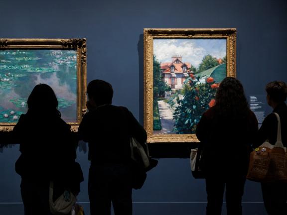 People with their backs to the frame look at two framed Impressionist paintings that hang on a dark wall.