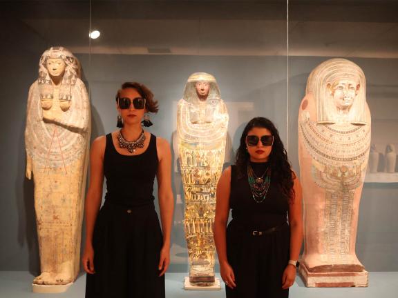 Two people wearing all-black tank tops and sunglasses stand with arms at their side in front of a display case full of Egyptian sarcophagi.
