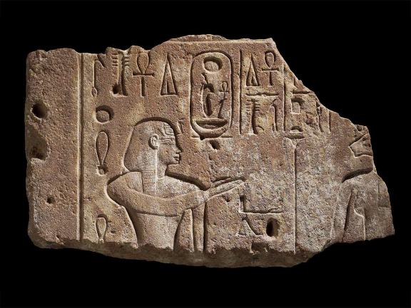 A quartzite carving in sunk relief showing Amenhotep III with a right hand raised toward before the lion-headed Sekhmet, who is barely seen due to the broken nature of the fragment. 