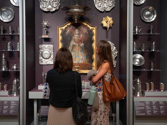 Two Museum visitors stand in front of case displays of richly ornamented silver objects surrounded by a painting of the virgin mary in a gold-tone frame