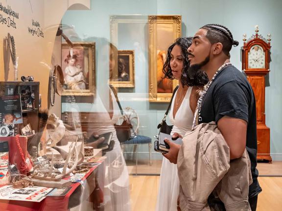 Profile view of two visitors looking at a display of an altar with Indigenous objects, ephemera, photos, and memorabilia, in an 18th-century art of the Americas gallery