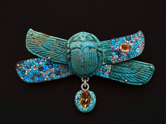 A turquoise scarab brooch with outstretched wings, adorned with blue and gold gemstones against a black background