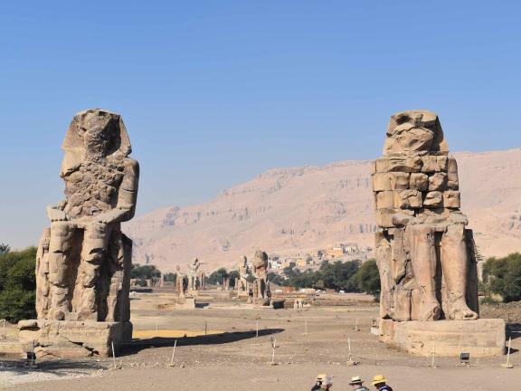 Two colossal stone statues, known as the Colossi of Memnon, stand in a desert landscape with distant mountains under a clear blue sky.