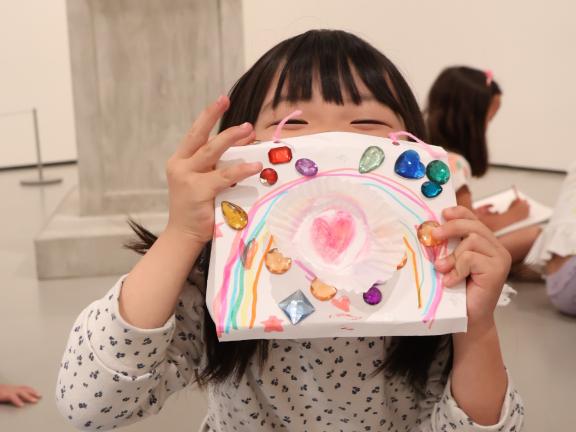 Young female student holds up artwork with gems in front of face (SAC)