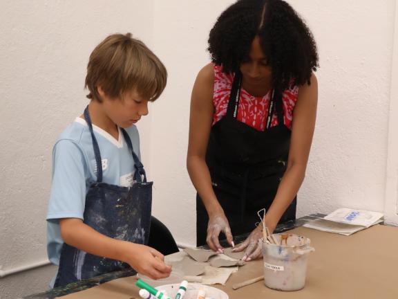 Studio Art Instructor assists young boy wearing a smock with clay project in classroom (SAC)