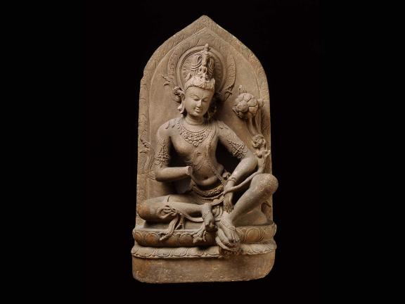 Intricate stone sculpture of a serene, seated figure in meditation, adorned with ornate jewelry and a crown, holding a lotus flower against a dark backdrop.