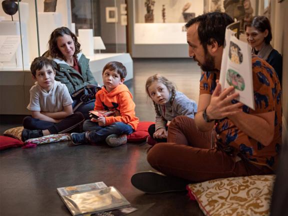 Three school-aged children and accompanying adults sit on a gallery floor and listen to a Museum educator read from a picture book