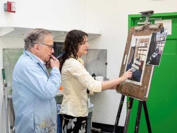 Studio Art Instructor critiques painting on easel from adult male student in blue smock in classroom (SAC) 