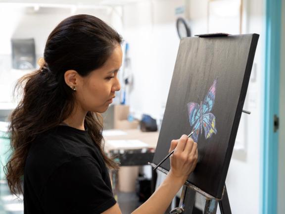 Adult female student paints butterfly on easel in classroom (SAC)