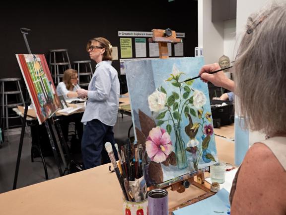 Adult student paints flowers on table top easel in classroom with students painting in background (SAC)