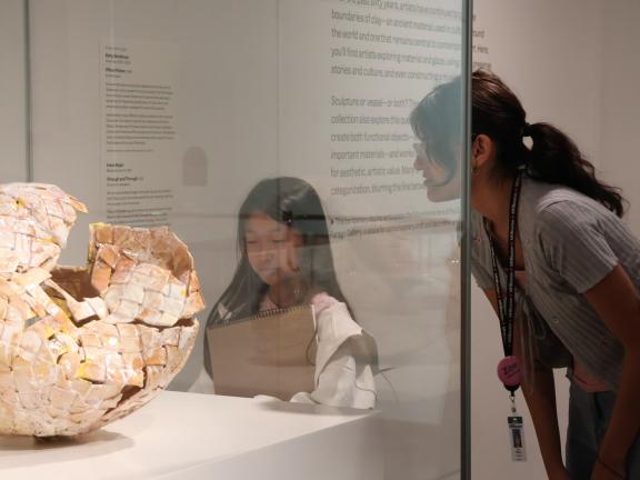 Studio Art Instructor looks at object in case with young female student holding a sketchbook (SAC)