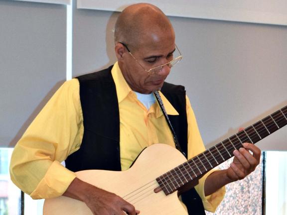 A man in a yellow shirt and black vest plays acoustic guitar.