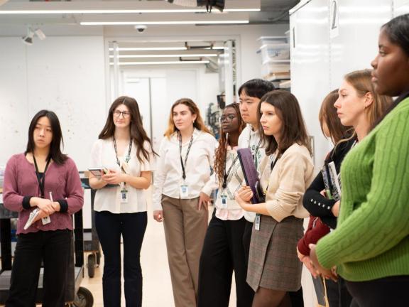 A group of young adults stand next to one another with notepads and appear to be listening to a speaker who is not in view