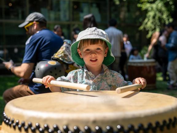 A child wearing a bucket hat plays a large traditional Japanese taiko drum outside on the Museum's Huntington Avenue Lawn