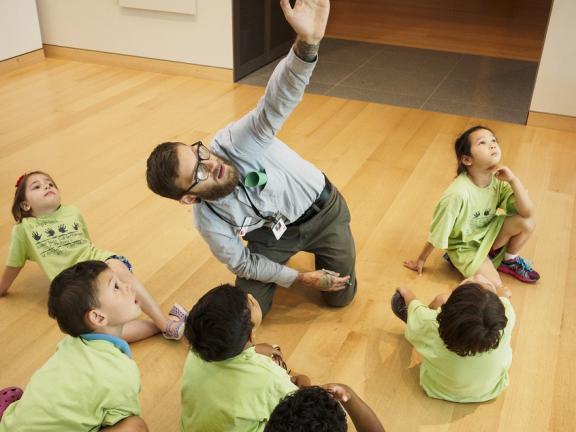 Teacher kneeling on gallery floor pointing up, with young kids in green shirts sitting around him